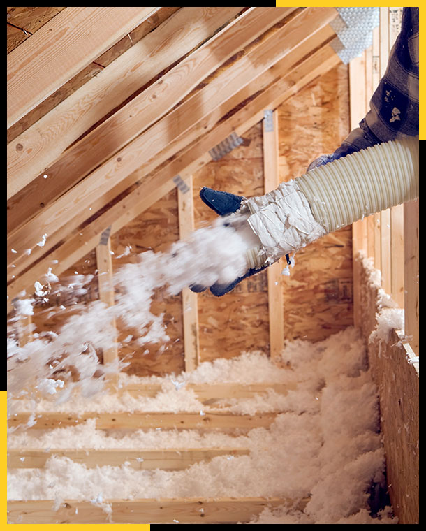 Worker spraying blown fiberglass insulation between attic trusses